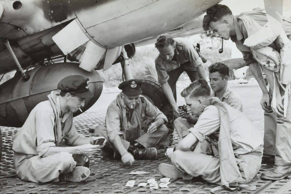 1943. A group of 77 Squadron RAAF pilots playing cards under a P-40 Kittyhawk fighter aircraft in New Guinea.